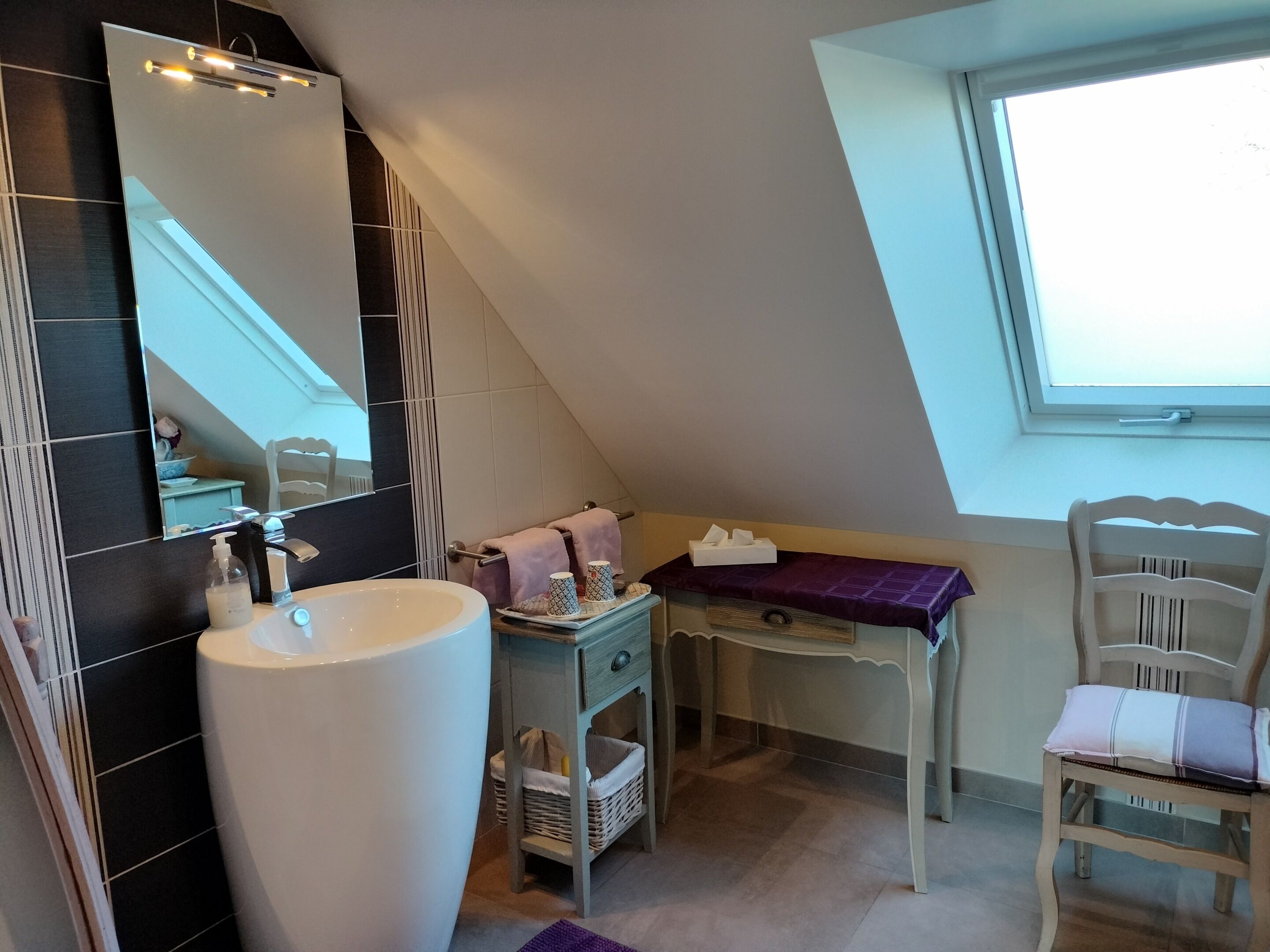 Une salle de bain avec un grand lavabo, un miroir, un porte-serviettes, une chaise et une fenêtre.