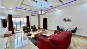 Una sala de estar moderna con un sofá rojo, una mesa de centro de madera y una mesa de comedor de vidrio. Hay un televisor montado en la pared, un candelabro y una ventana con cortinas rojas.