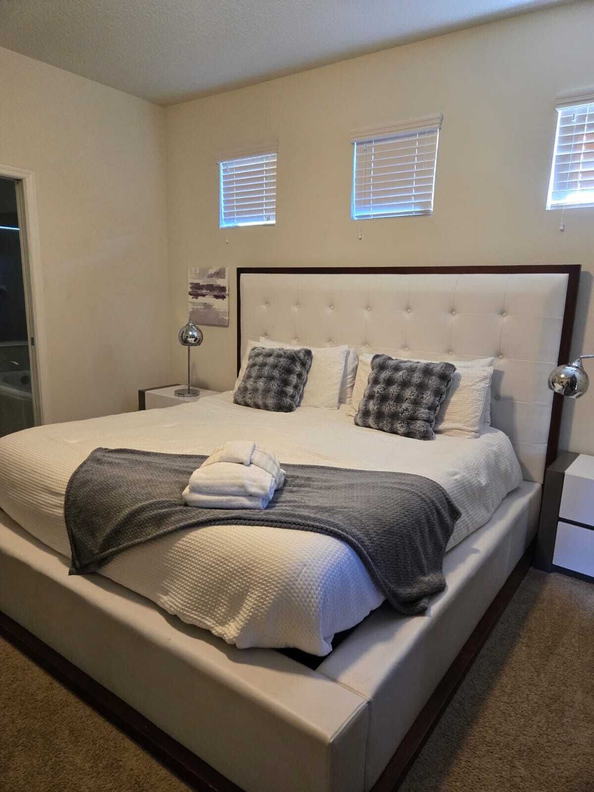 Una cama bien hecha con cabecera blanca, dos mesitas de noche y dos ventanas con persianas.
