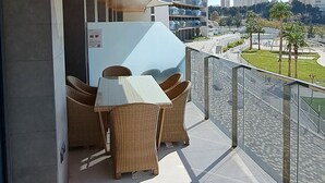 Un balcon doté de meubles en osier, d’une rambarde en verre et offrant une vue sur les bâtiments de la ville.