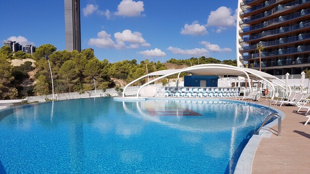 Une piscine d’un bleu éclatant, bordée d’une terrasse blanche, entourée d’arbres et d’un bâtiment moderne.