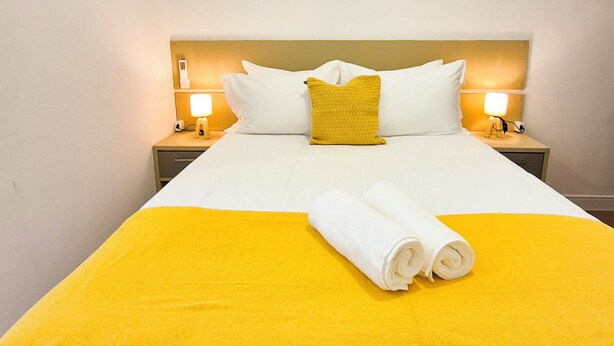 A hotel room with a bed, two bedside lamps, a yellow throw blanket, and two rolled towels. - 2 quartos à 400m praia de Copacabana (Rio de Janeiro)