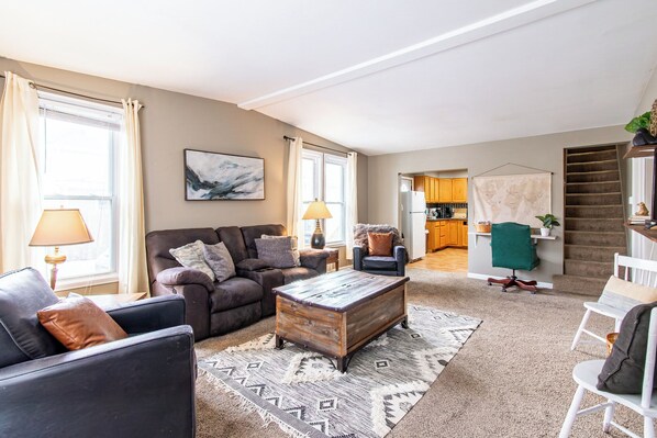A living room with a sofa, coffee table, and chairs, a kitchen in the background, and a staircase. - 3BR I Sleeps 8, Queen x 4, 5 minutes to Downtown (Fargo)
