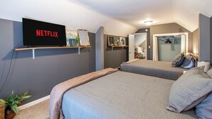 Une chambre avec deux lits, un téléviseur affichant le logo « NETFLIX », une étagère avec des livres et une plante, et une porte donnant sur une autre pièce.