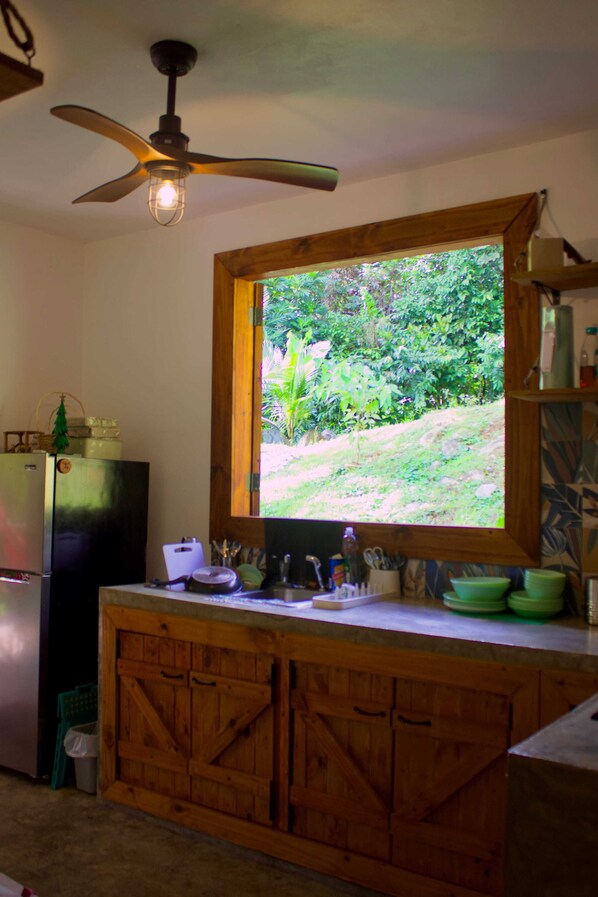 A kitchen with a wooden cabinet, a refrigerator, a window with a view of greenery, and a ceiling fan. - Finca Ki’ • Mar • Hammock • El Yunque Region • Ceiba Ferry • B&B (Naguabo)