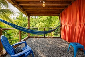 A hammock, blue chairs, and a red curtain in a covered outdoor area with a view of greenery. - Finca Ki’ • Mar • Hammock • El Yunque Region • Ceiba Ferry • B&B (Naguabo)