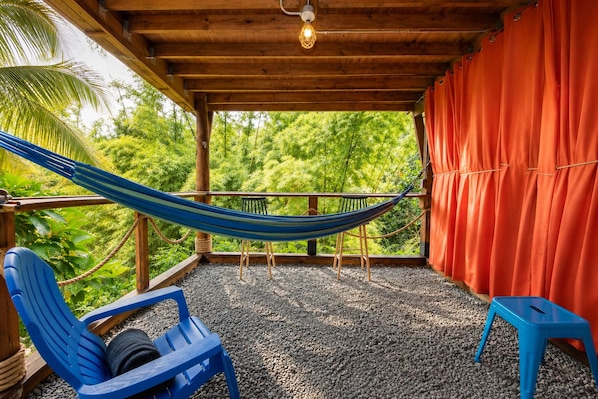 A hammock, blue chairs, and a red curtain in a covered outdoor area with a view of greenery. - Finca Ki’ • Mar • Hammock • El Yunque Region • Ceiba Ferry • B&B (Naguabo)