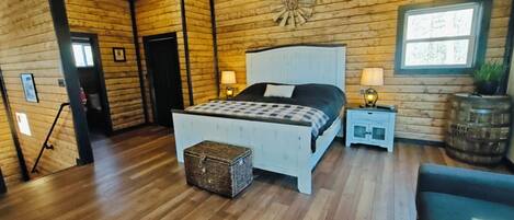 Une chambre avec un plafond en bois, un lit recouvert d’une couverture à carreaux, deux tables de chevet avec des lampes, un plancher en bois et une fenêtre donnant sur les arbres.