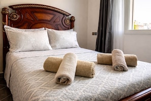 Una cama bien hecha con una colcha estampada, dos toallas enrolladas y un cabecero de madera.