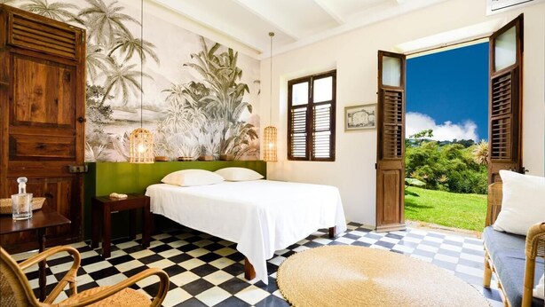Un dormitorio con piso a cuadros, cama con ropa blanca, armario de madera, una silla de mimbre y vista a la vegetación a través de contraventanas abiertas.