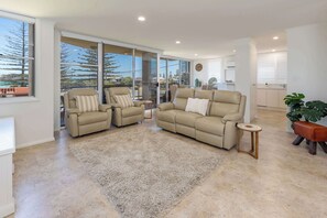 A spacious living area with a beige sofa set, a small round table, and a large window offering a view of the outdoors. - Beach Street Oasis - Central Lake Views - Tuncurry (Tuncurry)