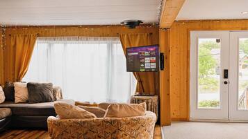 A cozy living room with a sofa, patterned cushions, a flat-screen TV mounted on the wall, and a wooden door leading outside.