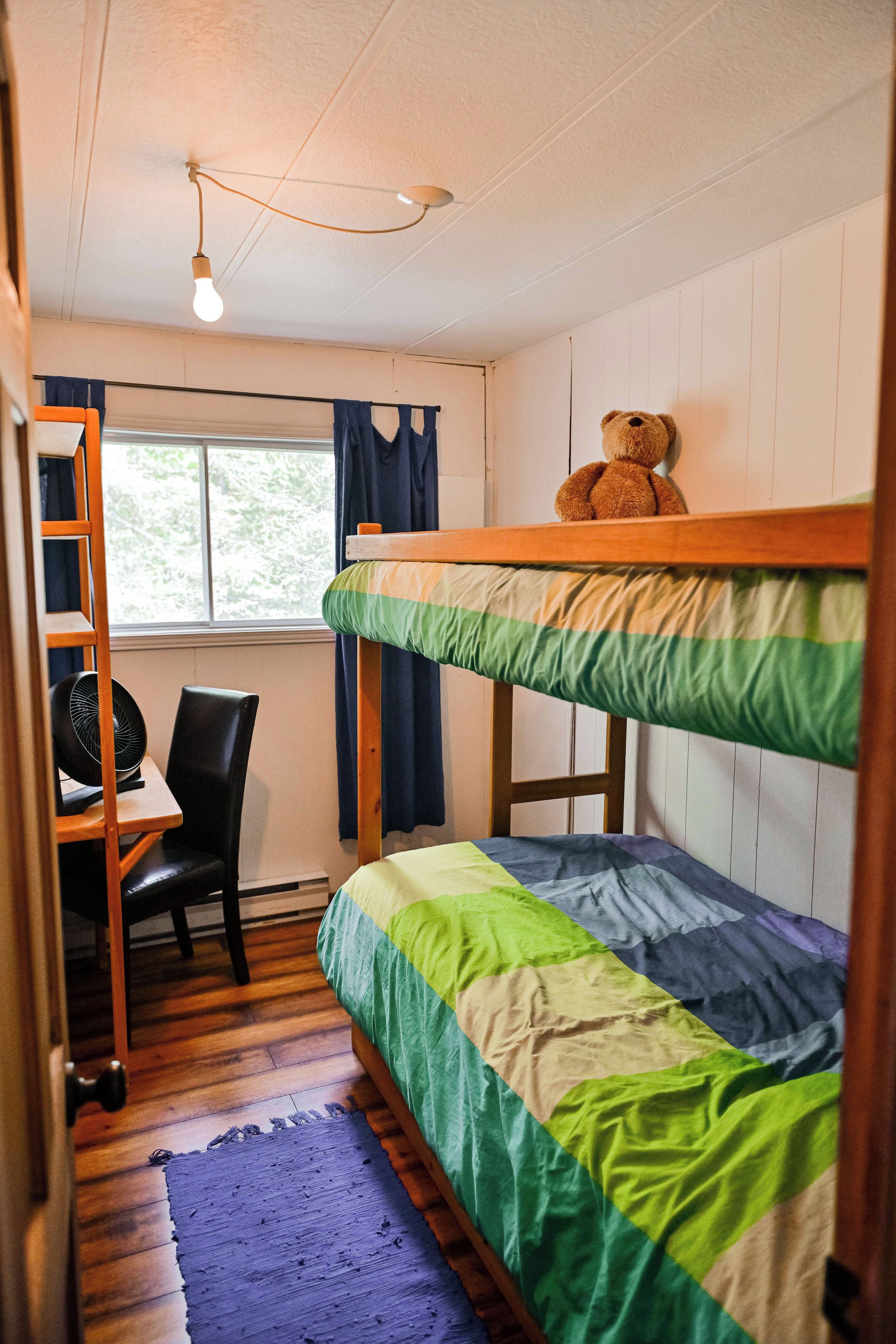 A bunk bed room with a desk and chair, a teddy bear on the top bunk, and a window with curtains.