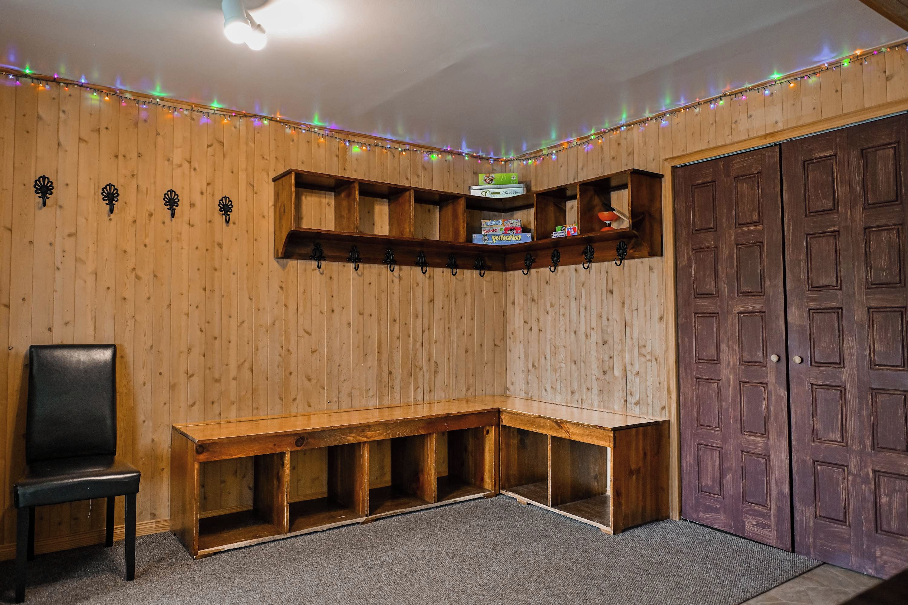 Quarto com paredes de madeira, um banco, uma porta e prateleiras com objetos.