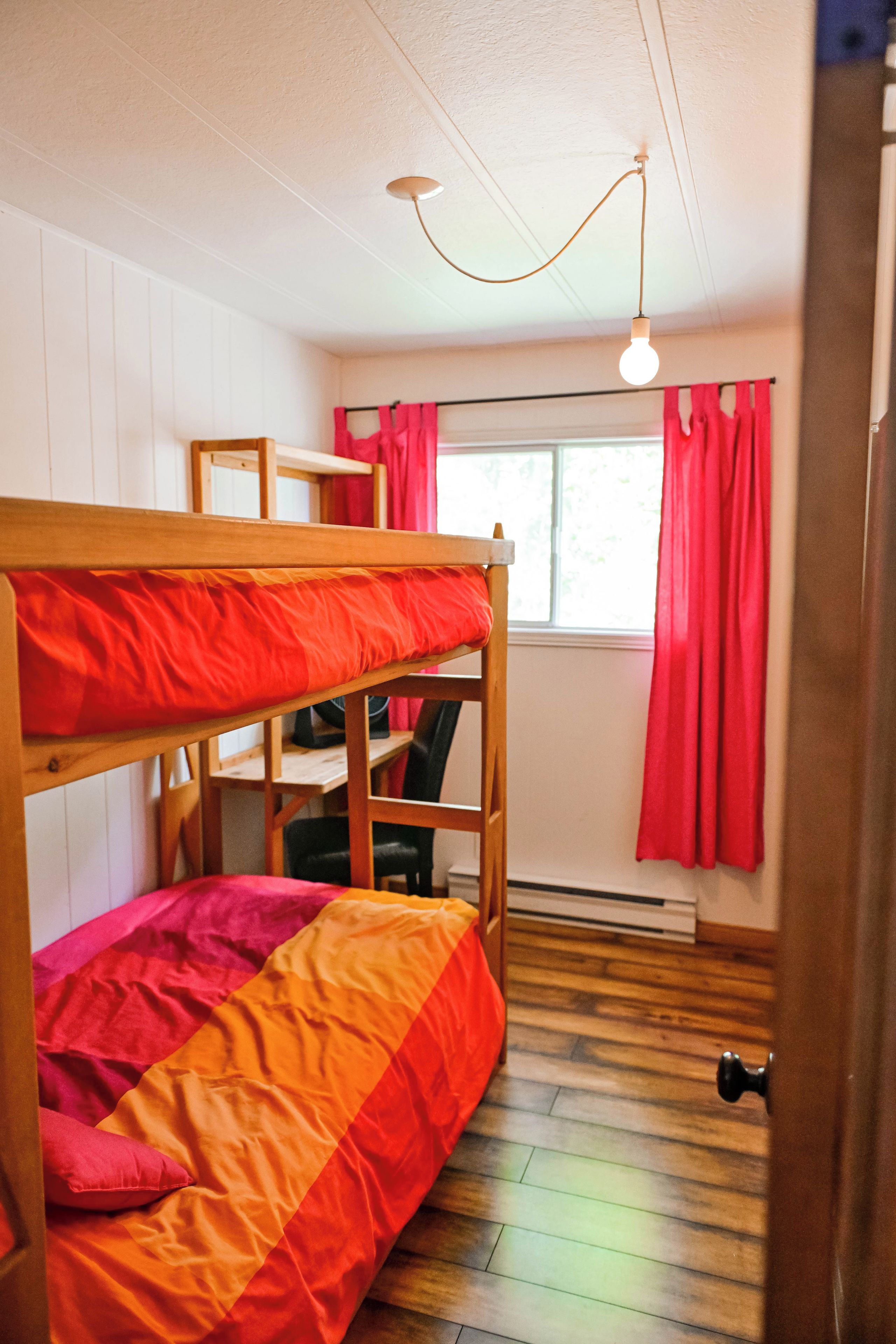 Uma cama beliche com colcha vermelha e laranja, uma escrivaninha com cadeira, uma janela com cortinas vermelhas e uma lâmpada pendente.