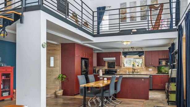 A modern kitchen with a central island, a dining area with a wooden table and chairs, and a staircase leading to an upper level. - L'Ecomaison - Luminous & Open Space (Rouyn-Noranda)
