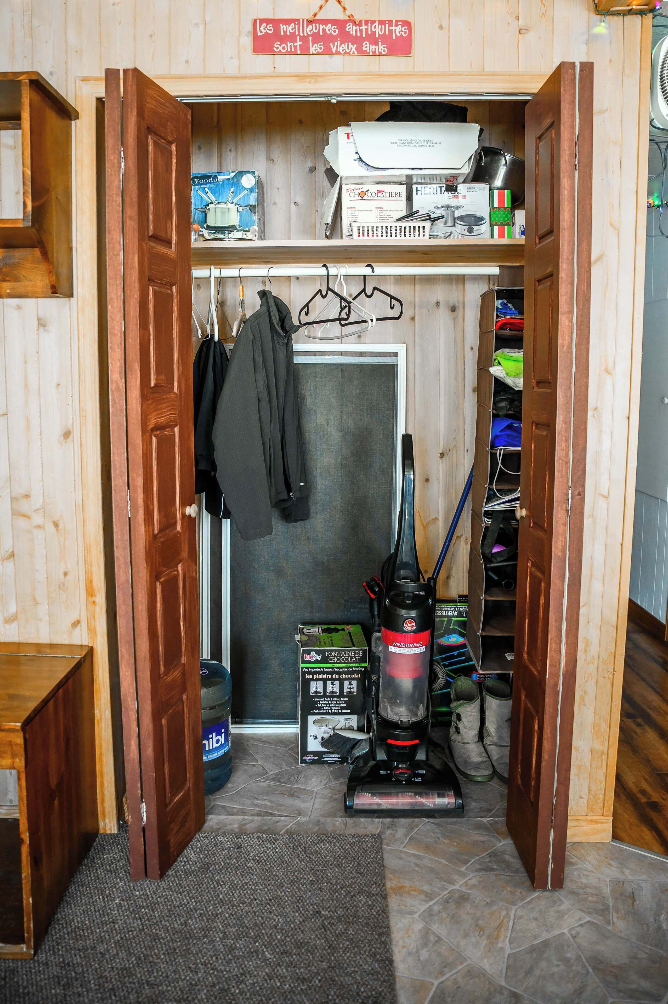 A wooden room with a door open, revealing a closet with hanging clothes, a vacuum cleaner, and various items on shelves.