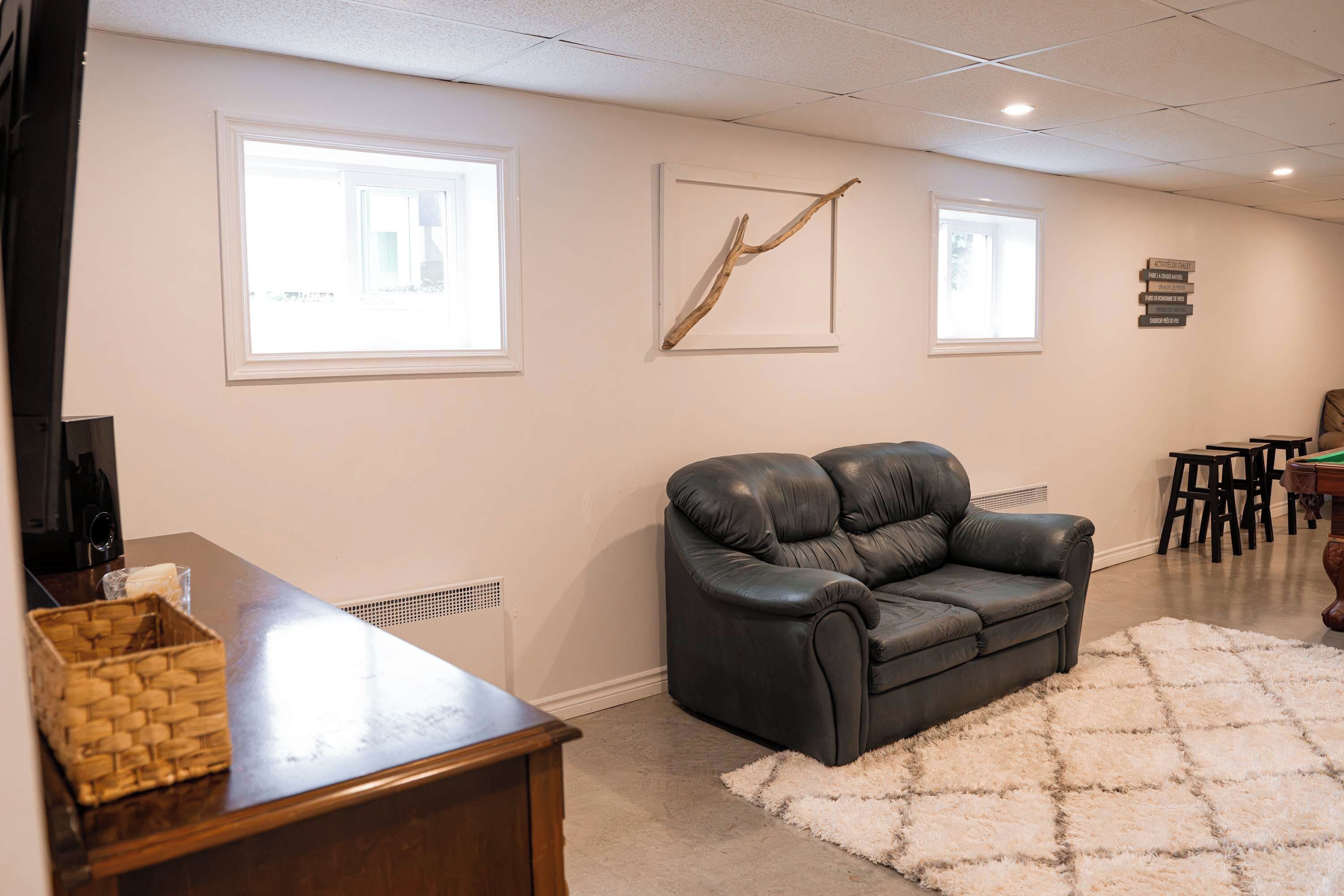 A living room with a black leather sofa, a wooden coffee table, and a white shag rug. There are two windows with blinds, a wall-mounted artwork, and a small table with chairs.