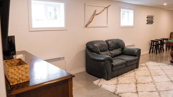 A living room with a black leather sofa, a wooden coffee table, and a white shag rug. There are two windows with blinds, a wall-mounted artwork, and a small table with chairs.