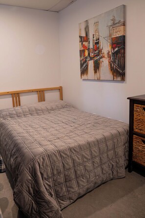 Quarto com cama, cabeceira, quadro na parede e um pequeno armário de madeira.