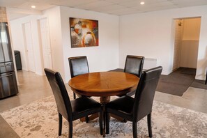 Uma sala de jantar com mesa redonda de madeira cercada por quatro cadeiras pretas, uma cozinha moderna com eletrodomésticos de aço inoxidável e uma pintura abstrata contemporânea na parede.