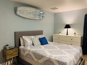 Un dormitorio con cama, mesita de noche, cómoda, lámpara y una tabla de surf en la pared.