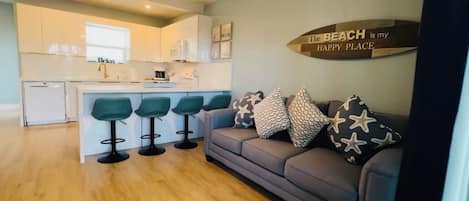 A modern kitchen with white cabinets and a central island, a grey sofa with star-patterned cushions, and a wooden sign on the wall.