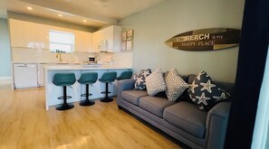 A modern kitchen with white cabinets and a central island, a grey sofa with star-patterned cushions, and a wooden sign on the wall. - Beach House: Turtle’s Cove #3-Full Kitchen, Washer and Dryer, 1st Floor (Melbourne Beach)