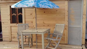 Un espace extérieur aménagé avec des meubles en bois, comprenant une table et deux chaises, un parasol aux motifs bleus et blancs, et une porte à volets fermés.