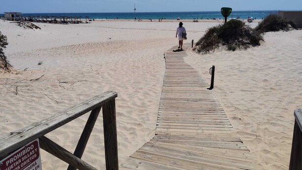 Une promenade en bois menant à une plage, avec un panneau indiquant qu’il est interdit d’y apporter certains objets.