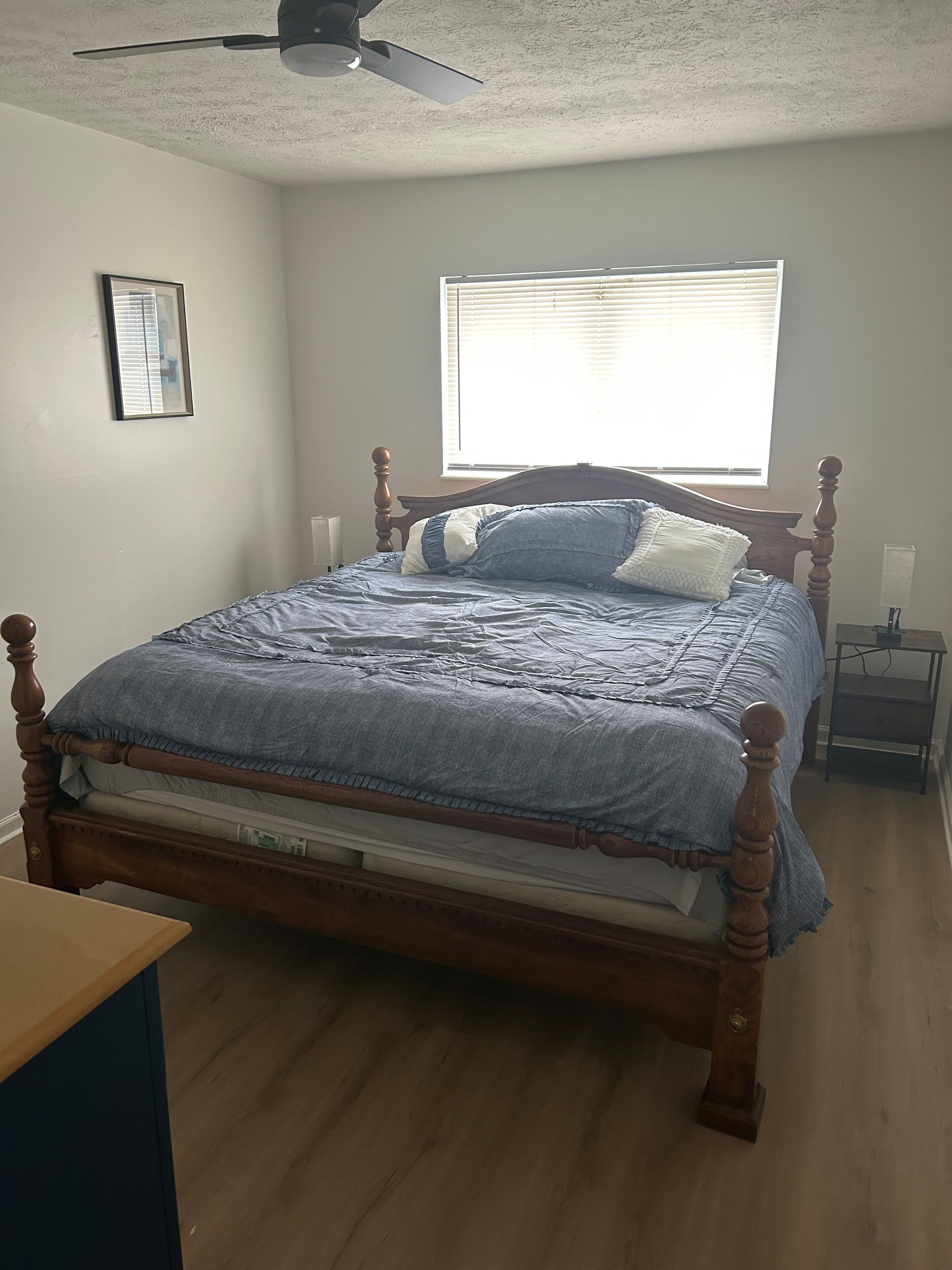 A bedroom with a wooden bed, a ceiling fan, a window with blinds, and a bedside table.