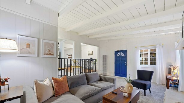 Una sala de estar con un sofá gris, una mesa de centro de madera y un sillón azul. Se ve una escalera que sube a otro nivel, una puerta azul y una ventana con cortinas blancas.
