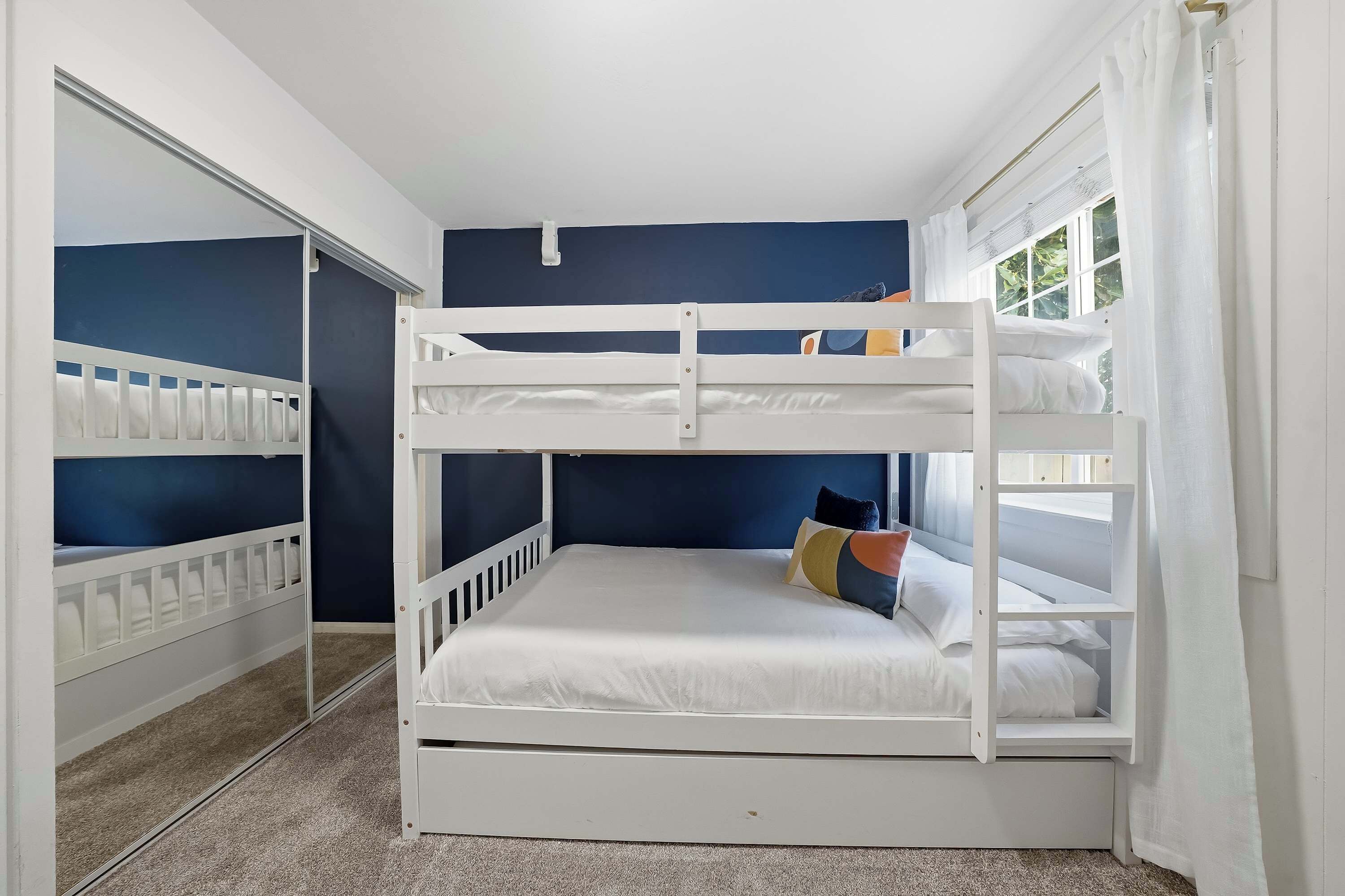 A bunk bed room with a lower bunk, a window with curtains, and a door with a mirror.