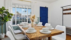 Un comedor con una mesa de madera dispuesta para cuatro, sillas blancas, un jarrón con plumas de pampa y una puerta azul.