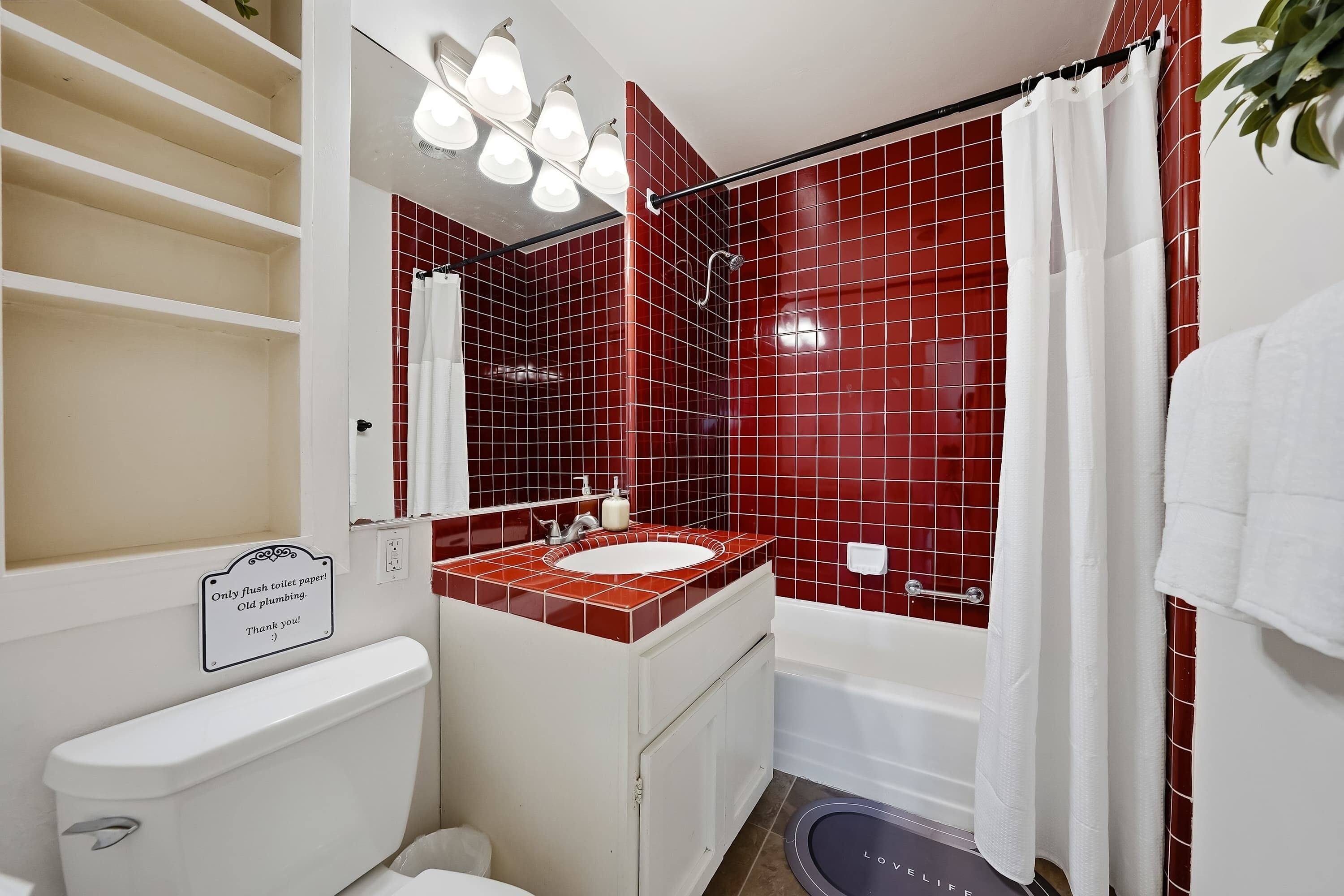 Een badkamer met een douche met rode tegels, een witte wastafel en een toilet. Boven het toilet staat een bord met de tekst: 'Alleen toiletpapier doorspoelen, Oude leidingen. Bedankt!'
