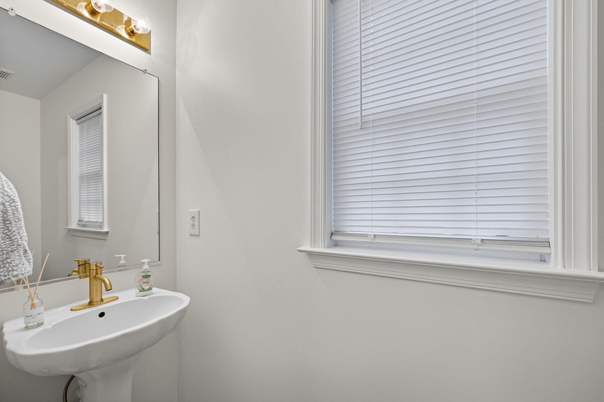 Een badkamer met een wit wastafelblad, een gouden kraan en een raam met witte luxaflex.
