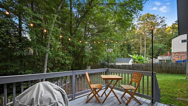 Une terrasse en bois avec deux chaises en bois et un couvercle de barbecue, entourée d’arbres et illuminée par une guirlande lumineuse.