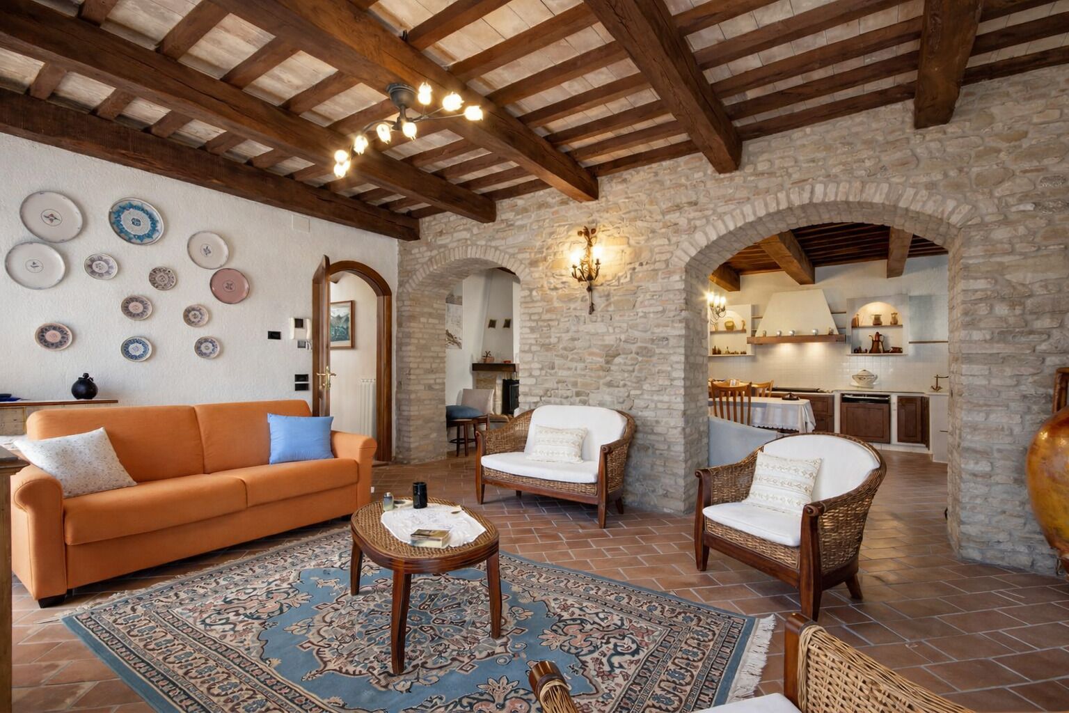 Un'ampia zona giorno con parete in pietra, travi in legno a soffitto e zona pranzo con cucina sullo sfondo.