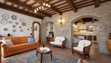 Un'ampia zona giorno con parete in pietra, travi in legno a soffitto e zona pranzo con cucina sullo sfondo.