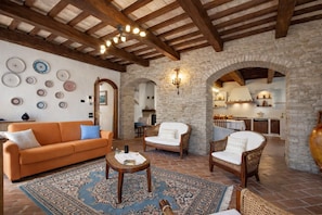 A spacious living area with a stone wall, wooden ceiling beams, and a dining area with a kitchen in the background. - Villa Angolo Fiorito (Marche)