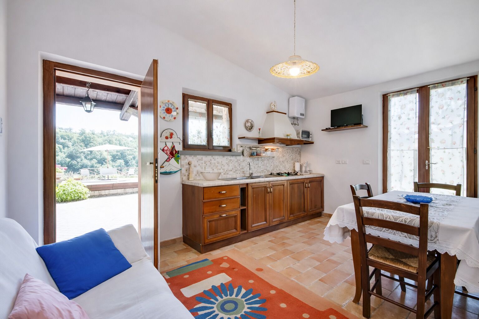 A compact kitchen with wooden cabinets, a dining area with a table and chairs, and a view through an open door to an outdoor area.