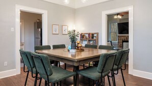 A dining room with a large wooden table, six green upholstered chairs, a vase with flowers, and a bookshelf with books and decorative items. - The Mooney House - Hot Tub, Grill, Screen Porch, Near Downtown (Hot Springs)