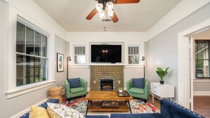 A living room with a fireplace, green armchairs, a sofa, and a flat-screen TV. - The Mooney House - Hot Tub, Grill, Screen Porch, Near Downtown (Hot Springs)