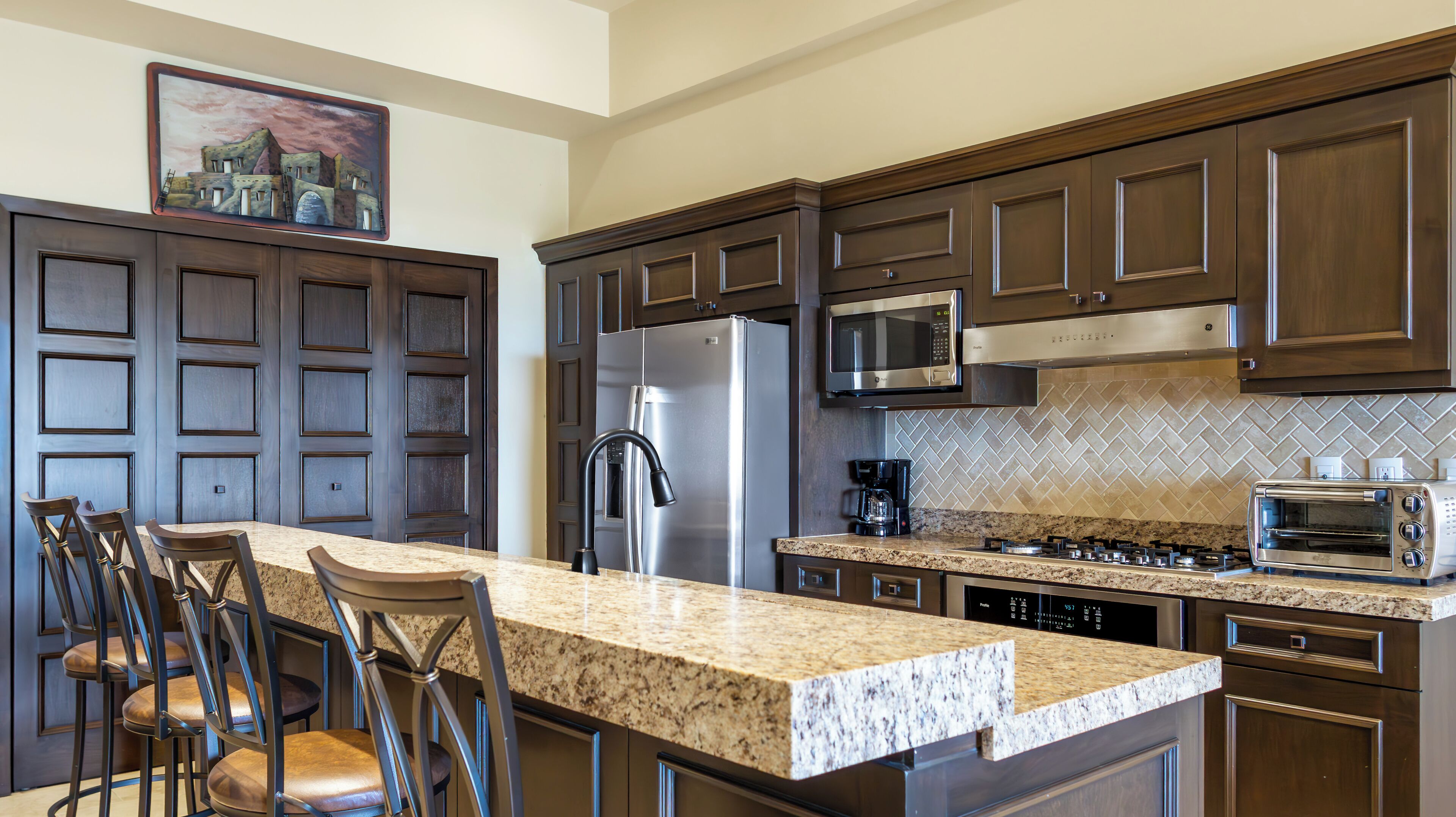 A modern kitchen with dark wood cabinets, a stainless steel refrigerator, a microwave, a gas stove, and a built-in oven. There are bar stools and a granite countertop.