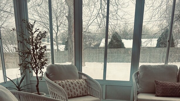 Un salón con ventanas amplio, sillones de mimbre, alfombra estampada y vista a un patio nevado.