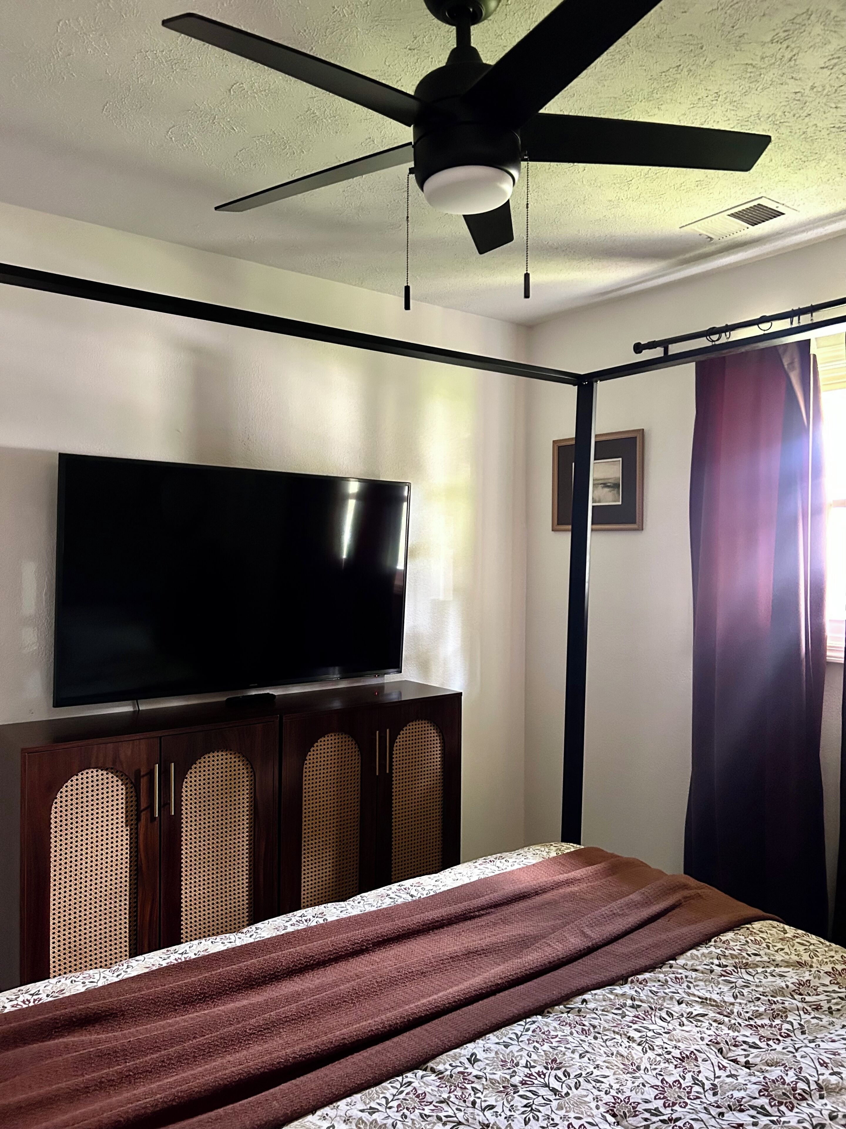 A bedroom with a bed, a flat-screen TV, a ceiling fan, and a window with curtains.
