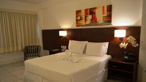 Habitación de hotel con una cama grande, dos lámparas de noche, un cabecero de madera, un cuadro sobre la cama y una silla junto a la ventana.