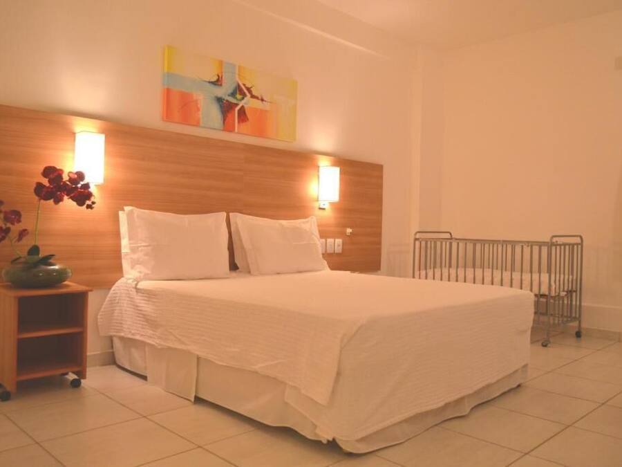 A hotel room with a large bed, two bedside lamps, a wooden headboard, a small bedside table with a vase of flowers, and a radiator.