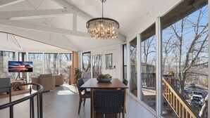 A spacious living room with a large window, a chandelier, and a dining area with a table and chairs. - Unplug, Unwind, Welcome to the Treehouse (Lake Ozark)