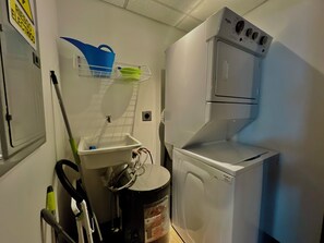 A compact laundry room with a front-loading washer and dryer, a sink with a mop, and a shelf with cleaning supplies. - Modern Central Apartment with Pool, Gym and Rooftop (San Salvador)